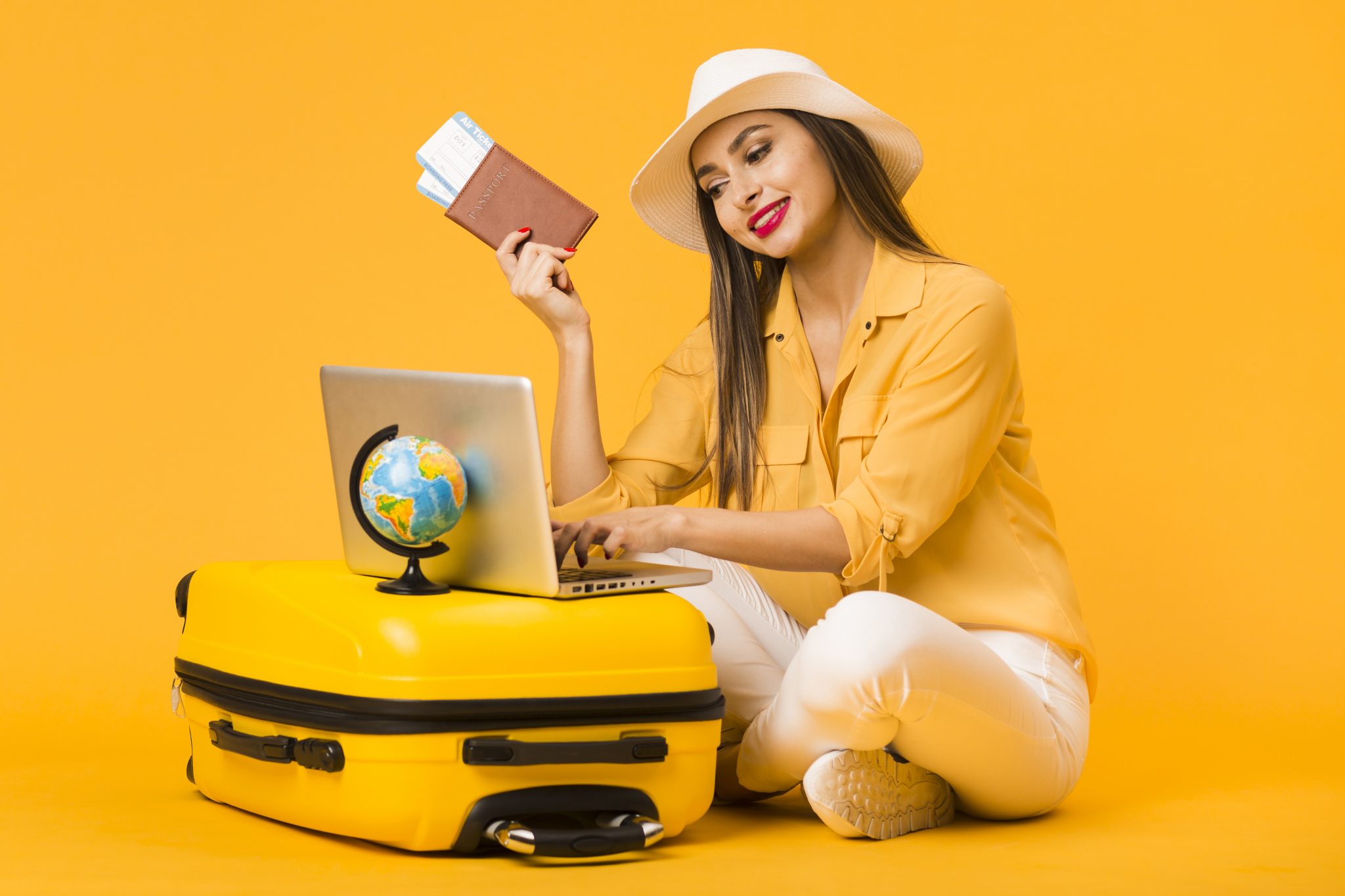  A woman wearing a summer hat and holding a passport and plane tickets is sitting on a yellow suitcase while using a laptop with a globe on it.